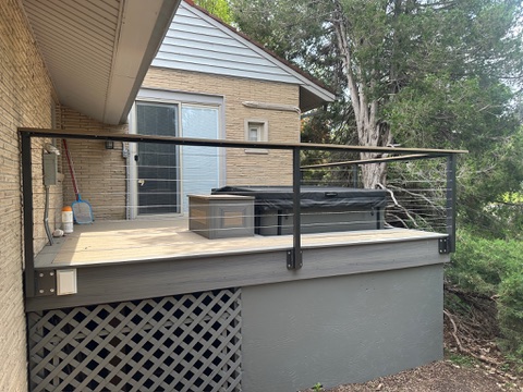 A deck in front of an A-frame style brick house with a window and glass door. Hanging attached to the railing is a broom a...